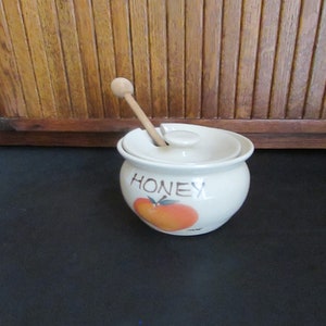 May include: A white ceramic honey pot with a lid and a wooden honey dipper. The pot has a brown and orange apple design and the word "HONEY" written on it.