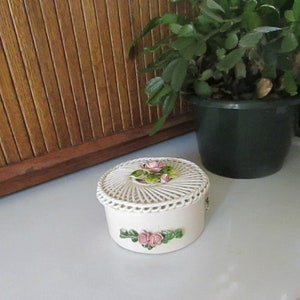 May include: A white ceramic trinket box with a woven lid and pink rose accents. The lid features a cluster of roses with green leaves. The box is decorated with a band of roses and leaves around the base.