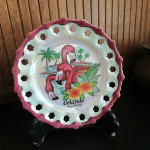 May include: A pink and white ceramic plate with a scalloped edge and cutouts around the rim. The plate features a painted image of two pink flamingos in a tropical setting with palm trees and flowers. The text "Orlando Florida" is printed on the plate.