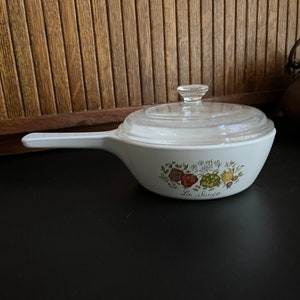 May include: White ceramic casserole dish with a glass lid. The dish has a floral design with a green, red, and yellow motif. The text "La Sauge" is printed on the bottom of the dish.