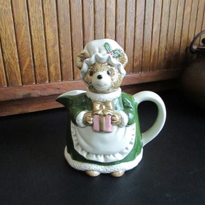 May include: A ceramic creamer in the shape of a bear wearing a green dress and a white bonnet. The bear is holding a pink and gold gift box.