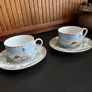 May include: Two white teacups with blue and white lighthouse designs on a nautical map pattern. The cups are on matching saucers.