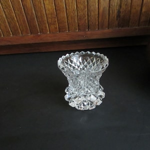 May include: A clear, cut-glass vase with a diamond pattern. The vase has a wide, scalloped rim and a flared base. The glass is thick and heavy, and the vase is likely vintage. The vase is sitting on a black surface.