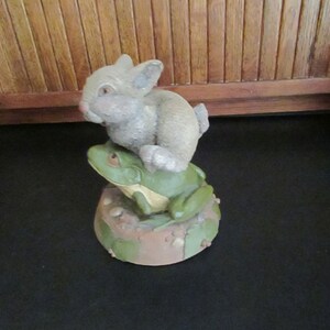 May include: A ceramic figurine of a green frog with a brown rabbit sitting on its back. The frog and rabbit are sitting on a brown base with green leaves and brown spots.