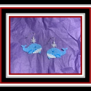 May include: A pair of blue whale earrings with white accents. The whales are made of beads and have a spray of water coming from their blowholes.