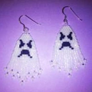 May include: A pair of handmade ghost earrings. Each earring features a white beaded ghost with a black pixelated face and silver-colored ear hooks. The earrings have a fringed bottom and are set against a purple background.