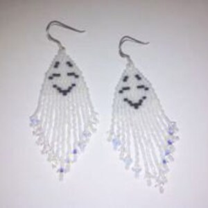 Puede incluir: Un par de pendientes de cuentas blancas con un diseño de cara sonriente. Los pendientes están hechos con cuentas de semillas blancas y tienen un gancho plateado.