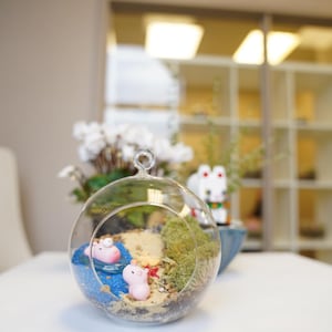 May include: A clear glass globe terrarium with a miniature beach scene. The terrarium contains sand, blue glitter, moss, and two small pink pig figurines.