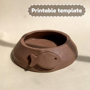 Puede incluir: Un cuenco de cerámica de color terracota con un diseño de pez. El cuenco es poco profundo y tiene un borde ancho. El diseño del pez está en relieve y tiene un pequeño agujero para un ojo.