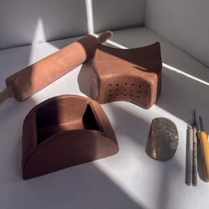 May include: A collection of terracotta-colored pottery tools and kitchenware. Includes a rolling pin, a perforated block, a curved container, a metal scraper, and several small tools with yellow and silver handles. The items are arranged on a white surface.
