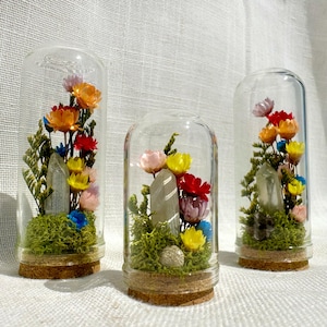 May include: Three glass cloches with cork bases, each containing a clear quartz crystal point nestled in green moss, surrounded by an arrangement of dried flowers in shades of orange, yellow, red, pink, and blue, displayed on a white fabric.