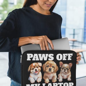 May include: Black laptop sleeve featuring three adorable puppy illustrations with the text "PAWS OFF MY LAPTOP" in white. The puppies are a mix of breeds, with brown, tan, and white fur. The sleeve is held by a person.