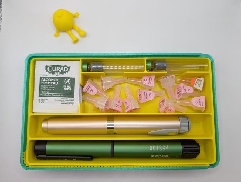 K&ouml;nnte beinhalten: Ein rechteckiges, blaugr&uuml;nes und gelbes Organizer-Tablett mit einem Curad-Alkoholtupfer, zwei Glasfl&auml;schchen, mehreren rosa Nadelspitzen, einem goldfarbenen Stift-Injektor und einem gr&uuml;nen Stift-Injektor mit der aufgedruckten Nummer 001894.