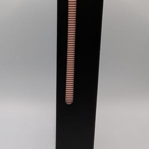 K&ouml;nnte beinhalten: Ein rechteckiges schwarzes elektronisches Ger&auml;t mit glatter, matter Oberfl&auml;che. An der Seite befindet sich ein vertikaler Schieberegler aus ros&eacute;goldenem Metall mit geriffelter Grifffl&auml;che und einem kleinen ovalen Knopf zur manuellen Steuerung.