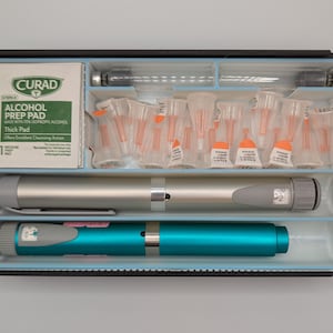 K&ouml;nnte beinhalten: Ein schwarzer medizinischer Aufbewahrungskoffer mit einem silbernen Injektionsstift, einem t&uuml;rkisfarbenen Injektionsstift, einem Curad-Alkohol-Vorbereitungspad und einer Reihe kleiner, klarer Nadelkappen mit orangefarbenen Spitzen in einem hellblauen Kunststoffeinsatz.