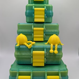 May include: A stack of four rectangular green storage containers with yellow latches, arranged in a pyramid shape. Two small, yellow, articulated figures with round bodies and dangling legs are seated on the second container from the bottom.