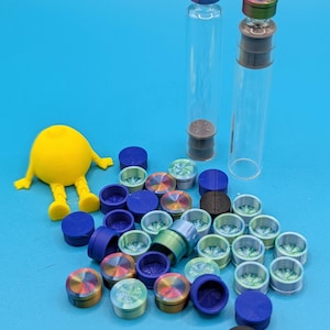 May include: A collection of colorful glass bottle stoppers with a variety of designs. The stoppers are arranged on a blue surface with two clear glass bottles and a yellow toy figure.