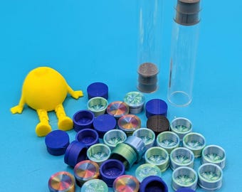May include: A collection of colorful glass bottle stoppers with a variety of designs. The stoppers are arranged on a blue surface with two clear glass bottles and a yellow toy figure.
