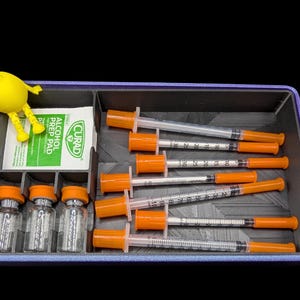 K&ouml;nnte beinhalten: Ein rechteckiges, violettes Aufbewahrungsfach mit sechs durchsichtigen Spritzen mit orangefarbenen Kappen, drei kleinen Glasfl&auml;schchen mit orangefarbenen Kappen und einem wei&szlig;en Curad-Alkoholtupfer. Eine kleine gelbe Gliederfigur sitzt am Rand.
