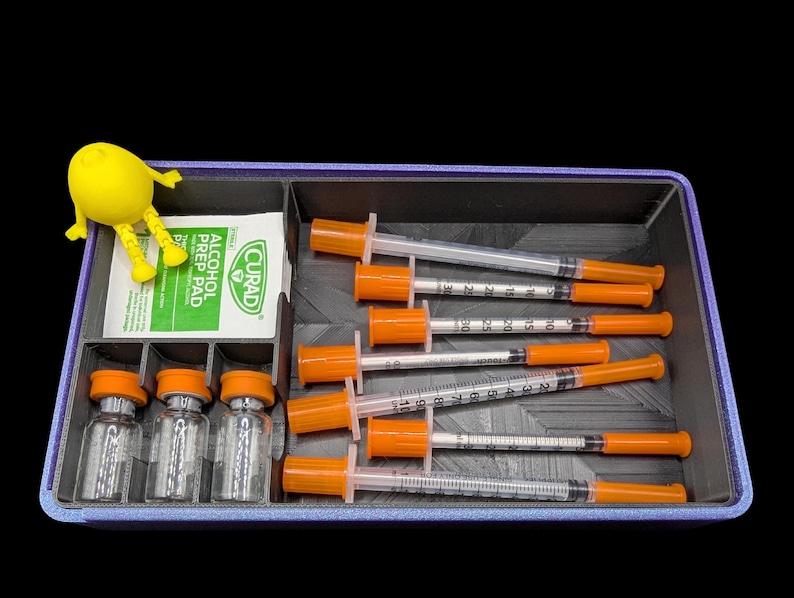 K&ouml;nnte beinhalten: Ein rechteckiges, violettes Aufbewahrungsfach mit sechs durchsichtigen Spritzen mit orangefarbenen Kappen, drei kleinen Glasfl&auml;schchen mit orangefarbenen Kappen und einem wei&szlig;en Curad-Alkoholtupfer. Eine kleine gelbe Gliederfigur sitzt am Rand.