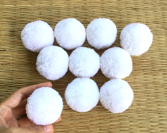 Può includere: Nove pompon bianchi disposti in cerchio su un tappetino di paglia intrecciata. Un pompon è tenuto da una mano.