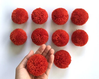 May include: Nine orange pom-poms arranged in a grid pattern on a white background. One pom-pom is being held in a hand.