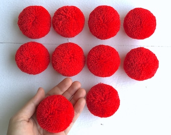 May include: Ten bright red pom poms, each about 5 cm in diameter, are arranged in a semi-circle on a white surface. One pom pom is being held in a hand.