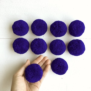 May include: Ten purple pom poms, approximately 2 inches in diameter, arranged in a grid pattern on a white surface. One pom pom is being held in a hand.