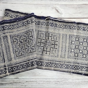 May include: A folded textile with a repeating geometric pattern in navy blue and cream. The design features cross-like shapes and floral motifs within rectangular borders. The fabric has a fringed edge and is displayed on a light wood surface.