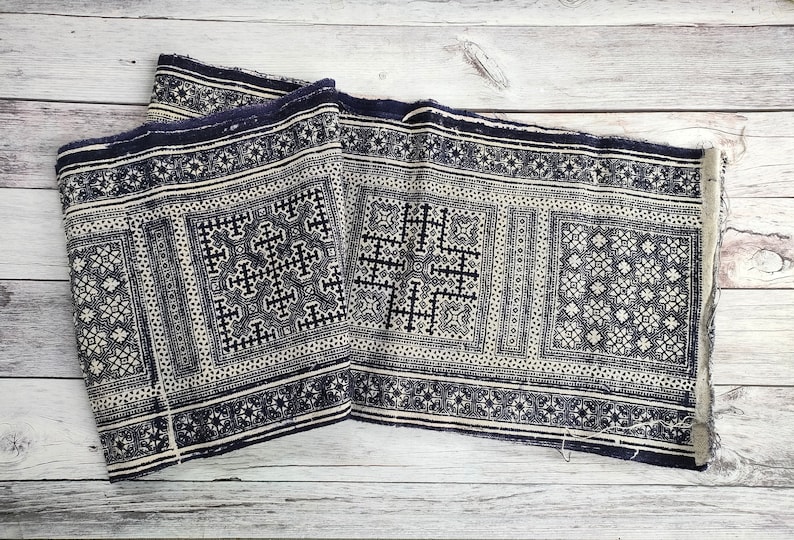 May include: A folded textile with a repeating geometric pattern in navy blue and cream. The design features cross-like shapes and floral motifs within rectangular borders. The fabric has a fringed edge and is displayed on a light wood surface.