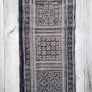 May include: A long, rectangular textile with a repeating geometric pattern in navy blue and off-white. The design features squares and cross-like shapes, bordered by vertical stripes. The fabric appears to be woven or embroidered.