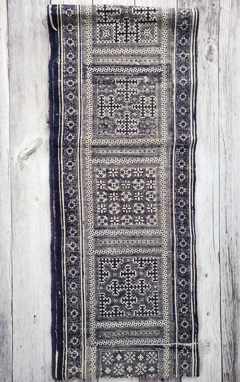 May include: A long, rectangular textile with a repeating geometric pattern in navy blue and off-white. The design features squares and cross-like shapes, bordered by vertical stripes. The fabric appears to be woven or embroidered.