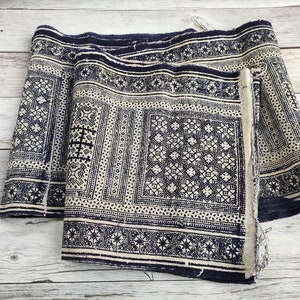 May include: Two rolls of fabric featuring a repeating geometric pattern in navy blue and off-white. The fabric has a detailed design with squares, stars, and other shapes. The edges of the fabric are frayed.