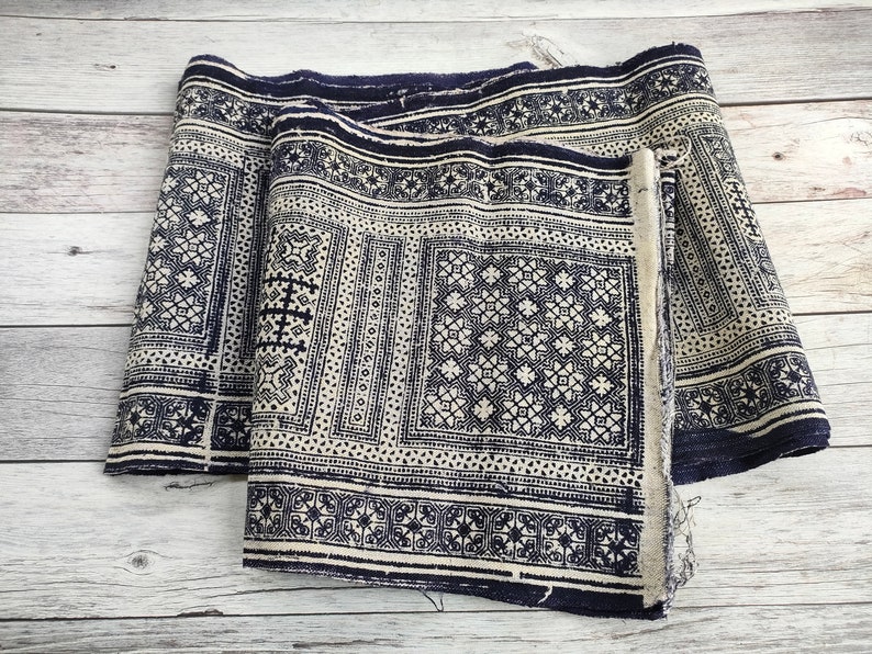 May include: Two rolls of fabric featuring a repeating geometric pattern in navy blue and off-white. The fabric has a detailed design with squares, stars, and other shapes. The edges of the fabric are frayed.