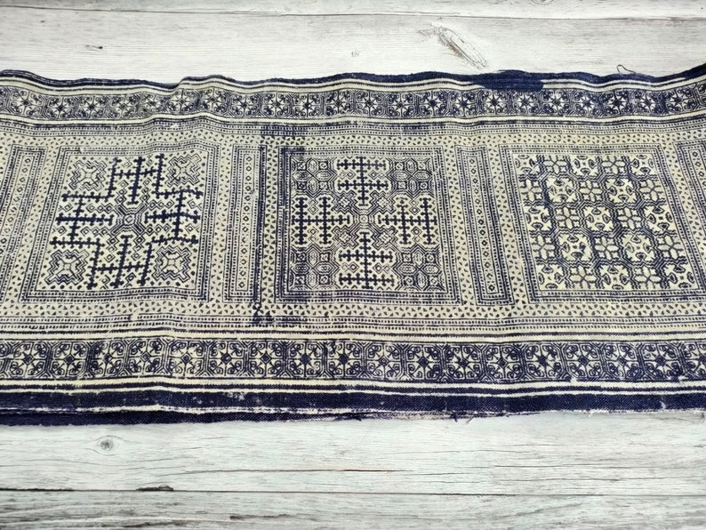 May include: A length of fabric displaying a repeating pattern in navy blue and off-white. The design incorporates geometric shapes, such as crosses and squares, within rectangular panels. The edges are finished with a navy blue border.