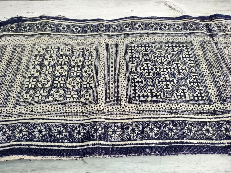 May include: A length of indigo and cream fabric with a detailed geometric design. The pattern includes repeating square and rectangular motifs with cross and floral elements. The fabric has a textured look and a dark indigo border, suitable for various craft projects.