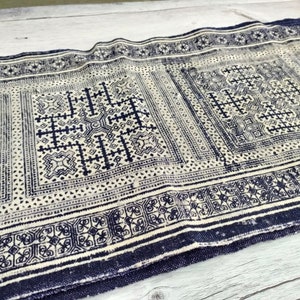 May include: A rectangular textile with a repeating geometric pattern in navy blue and off-white. The design features squares and rectangles with intricate cross and floral motifs, bordered by a dark blue edge. The fabric appears to be woven.