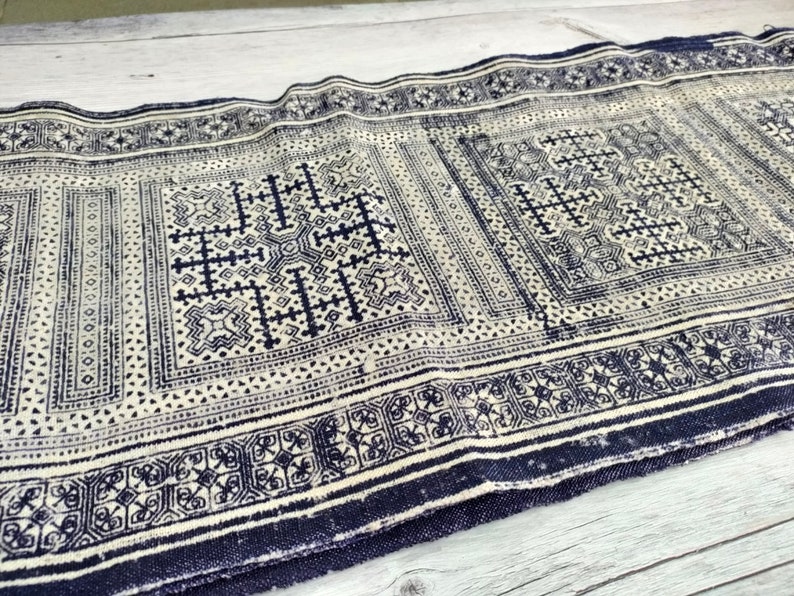 May include: A rectangular textile with a repeating geometric pattern in navy blue and off-white. The design features squares and rectangles with intricate cross and floral motifs, bordered by a dark blue edge. The fabric appears to be woven.