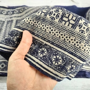 May include: Close-up of a folded textile with a repeating geometric pattern in navy blue and off-white. The fabric has a textured appearance, with detailed designs and borders. The textile is likely used for home decor or fashion.