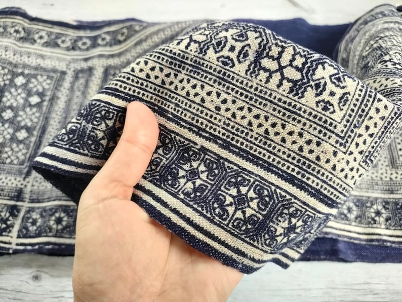 May include: Close-up of a folded textile with a repeating geometric pattern in navy blue and off-white. The fabric has a textured appearance, with detailed designs and borders. The textile is likely used for home decor or fashion.