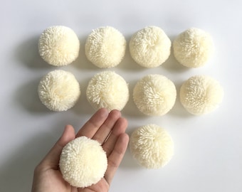 May include: Ten cream-colored fluffy pom poms, each about 2 inches in diameter, are arranged in a row on a white surface. One pom pom is held in a hand, showing the texture and size.