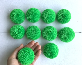 Può includere: Dieci pompon morbidi di colore verde brillante, ognuno di circa 5 cm di diametro, sono disposti in tre file su una superficie bianca. Un pompon è tenuto in una mano.