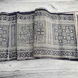 May include: A folded textile with a repeating geometric pattern in navy blue and cream. The design features squares, crosses, and star-like shapes. The fabric appears to be woven, with a slightly distressed edge.