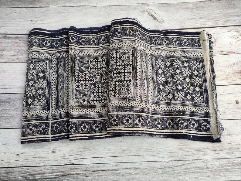 May include: A folded textile with a repeating geometric pattern in navy blue and cream. The design features squares, crosses, and star-like shapes. The fabric appears to be woven, with a slightly distressed edge.