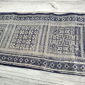 May include: A rectangular textile with a repeating geometric pattern in navy blue and off-white. The design features squares and borders with intricate cross and floral motifs. The fabric appears to be woven or printed.