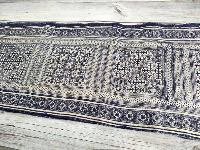 May include: A rectangular textile with a repeating geometric pattern in navy blue and off-white. The design features squares and borders with intricate cross and floral motifs. The fabric appears to be woven or printed.