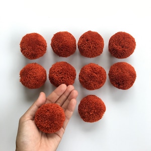 May include: Ten orange pom poms, each about 2 inches in diameter, are arranged in a grid pattern on a white surface. One pom pom is being held in a hand.