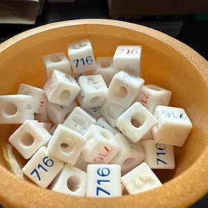 May include: A collection of small, white, square blocks with the number "716" printed on them in blue and red. The blocks are inside an orange bowl. Some blocks have a hole in the center.