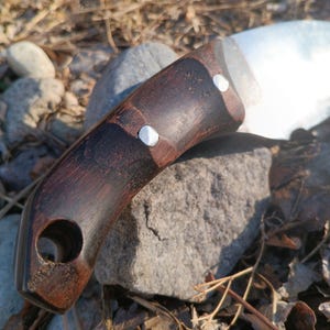 以下が含まれることがあります： 暗い木製のハンドルと銀色の刃を持つ手作りナイフ。ハンドルには銀色のリベットが2つと、下部に穴があります。