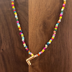 May include: A colorful beaded necklace with a gold-toned pendant. The necklace features small, round beads in various colors, including pink, yellow, blue, and green. The pendant is a geometric shape. The necklace is displayed on a wooden surface.